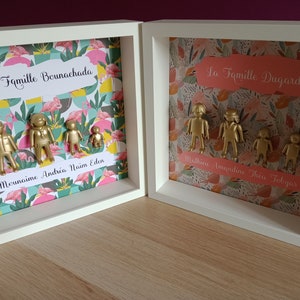 May include: Two white square shadow box frames containing gold-painted plastic figures arranged as families. The left frame features a flamingo pattern and the text La Famille Bounachada and Mounaime Andr&eacute;a Na&iuml;m Eden. The right frame has a floral pattern and the text La Famille Dugard and Mathieu Amandine Th&eacute;o Tobyas.