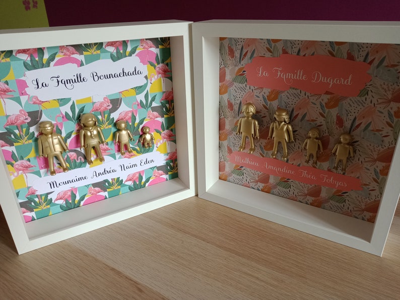 May include: Two white square shadow box frames containing gold-painted plastic figures arranged as families. The left frame features a flamingo pattern and the text La Famille Bounachada and Mounaime Andr&eacute;a Na&iuml;m Eden. The right frame has a floral pattern and the text La Famille Dugard and Mathieu Amandine Th&eacute;o Tobyas.