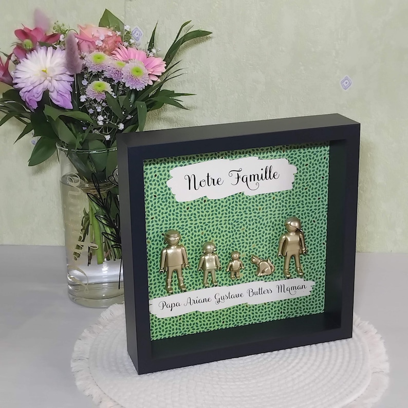 May include: A black shadow box frame containing a green patterned background with gold-colored toy figures of two adults, two children, and a cat. The text "Notre Famille" appears at the top, and "Papa Ariane Gustave Butters Maman" at the bottom.