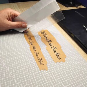 May include: Two tan adhesive vinyl decals with black cursive text. One decal reads La famille du Bonheur and the other reads Louise Mila Tiago Chlo&eacute; Jean Mael. A clear transfer sheet is being applied to the decals on a gray grid cutting mat.