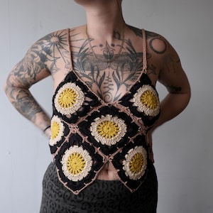 Könnte beinhalten: Handgefertigtes Häkeltop mit floralem Design. Das Oberteil besteht aus schwarzen, cremefarbenen und gelben Quadraten mit einem braunen Träger. Dieses einzigartige Kleidungsstück ist ein tragbares Kunstwerk, ideal für einen Boho- oder alternativen Stil.