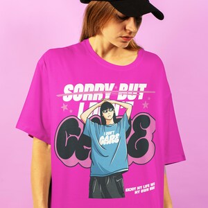 May include: A pink t-shirt featuring a graphic print of a woman wearing sunglasses and the text "Sorry But I Don't Care" in white lettering. The text is crossed out with a pink line. The woman is wearing a blue shirt with the text "I Don't Care" in white lettering. The graphic is surrounded by black and pink clouds. The text "Enjoy My Life WI My Own RuI" is printed in white lettering at the bottom of the shirt.