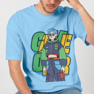 May include: Light blue t-shirt featuring a graphic print of a cartoon character wearing a blue, green and black jacket with the text "GAME OVER" on the front. The character has blue hair and is wearing blue jeans with a red pocket.