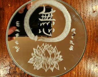 以下が含まれることがあります： 中央に蓮の花、手描きの円、日本語の漢字が白くエッチングされた円形の鏡。鏡面には暗い長方形の物体が映り込んでおり、側面には白い文字が描かれています。鏡の表面には、繊細な白い線で描かれた植物や幾何学的な模様が施されています。