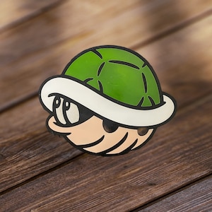 May include: A green and white cartoon character with a shell on its head. The character has large eyes and a small mouth. The shell is green with black lines and a white rim.