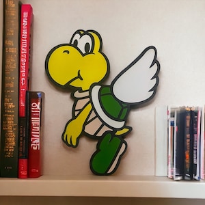 May include: A yellow and green cartoon character with wings, a shell, and a pink and white striped shirt. The character is standing on a shelf with books behind it.