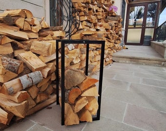 May include: A black metal firewood rack filled with stacked pieces of firewood. The rack is sitting on a stone patio in front of a stone building.