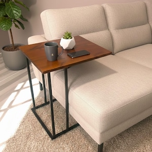 May include: A brown wooden side table with a black metal frame is positioned next to a beige sofa. A gray mug, a small white geometric planter with a succulent, and a black smartphone rest on the table's surface. A potted plant is visible in the background.