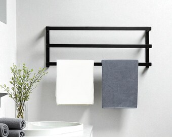 May include: A black metal towel rack with three horizontal bars, holding a white and a gray towel. The rack is mounted on a white wall above a white countertop with a sink and rolled towels.