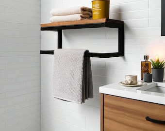 May include: A modern bathroom shelf with a black metal frame and a wooden top. The shelf holds folded beige towels and a yellow container with the word "Hygge". A gray towel hangs below. The bathroom has white tile walls and a wooden vanity.