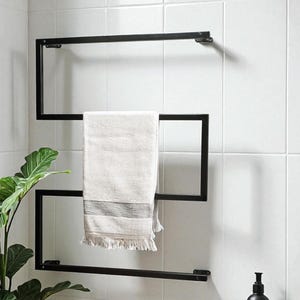 Può includere: Un portasciugamani in metallo nero dal design geometrico, che tiene un asciugamano bianco con strisce grigie e frange. Il portasciugamani è montato su una parete piastrellata bianca, con una pianta e accessori da bagno sullo sfondo.