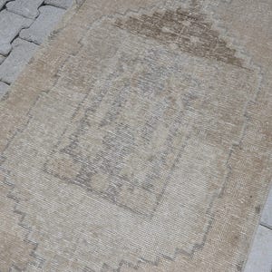 May include: A rectangular woven rug featuring a muted beige and light brown colour palette. The design includes a central geometric medallion with a stepped outline and a faded, distressed texture throughout the surface of the textile piece.