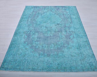 May include: A rectangular, distressed-look rug in shades of teal and blue. The rug features a central medallion design and a border with a faded, vintage appearance. The rug is placed on a light-colored wooden floor.