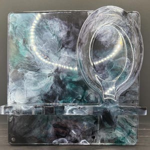 May include: A clear resin soap dish with a built-in soap holder. The resin is swirled with shades of teal, black, and white. The soap dish is rectangular with a curved soap holder attached. The dish sits on a dark wooden surface.
