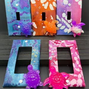 May include: A collection of decorative light switch covers with colorful, abstract designs. The covers feature shades of blue, orange, and pink, adorned with small, translucent turtle embellishments. The designs include patterns of white and other colors.
