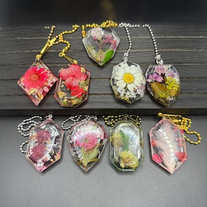 May include: A collection of eight resin pendants, each encasing dried flowers in various shapes. The pendants feature gold and silver chains. The flowers are in shades of red, pink, white, and yellow, with gold leaf accents.