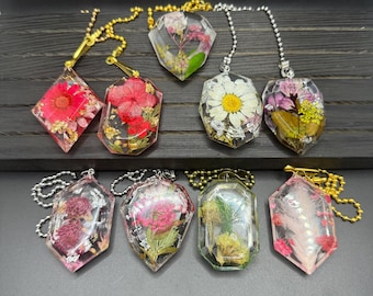 May include: A collection of eight resin pendants, each encasing dried flowers in various shapes. The pendants feature gold and silver chains. The flowers are in shades of red, pink, white, and yellow, with gold leaf accents.