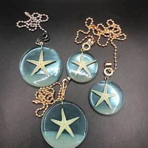 May include: Four decorative ceiling fan pull chains. Each chain features a clear, round pendant with a starfish encased in blue resin. The chains are silver and gold-toned, with varying pendant sizes.