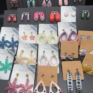 May include: A collection of various resin earrings displayed on individual cards. The assortment includes floral, teardrop, geometric, and moon phase shapes in diverse colors like pink, blue, green, and glitter finishes, all featuring metal earring posts.