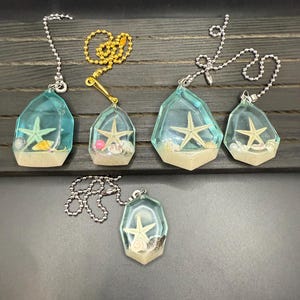 May include: A collection of six decorative ceiling fan pull chains. Each chain features a clear, geometric pendant with a starfish and sand-like material encased within. The pendants are connected to metal chains of varying colors.