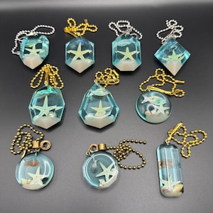 May include: A collection of ten resin keychains featuring miniature starfish, seashells, and small fish figures encased in clear blue resin. Each piece includes a metal ball chain in either silver or gold tone, with some shapes being geometric, circular, or rectangular.