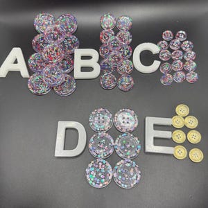 May include: A collection of round buttons arranged in groups labeled A through E. Groups A, B, C, and D feature clear buttons with embedded colorful star-shaped glitter. Group E consists of smaller, solid gold-colored buttons with four holes.