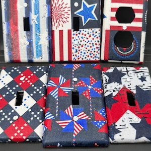 May include: A collection of decorative light switch covers with patriotic designs. The covers feature patterns of stars, stripes, and pinwheels in red, white, and blue. Some have a single switch opening, while others have a double outlet.