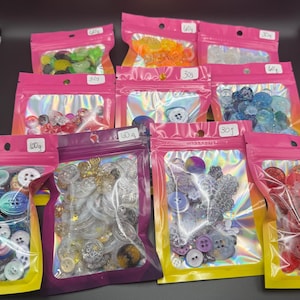 May include: Assortment of colorful buttons in various shapes and sizes, packaged in pink and holographic zip-lock bags. Weights are labeled in grams and ounces. The buttons include clear, iridescent, and solid colors, perfect for crafts and sewing projects.
