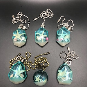 May include: A collection of six ocean-themed ceiling fan pull chains. Each chain features a clear, geometric pendant with a starfish, shells, and sand encased in resin. The pendants are attached to silver or brass-colored ball chains.