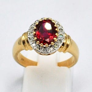 Może przedstawiać: Złoty pierścionek z dużym, owalnym, głęboko czerwonym kamieniem, otoczonym mniejszymi, przezroczystymi kamieniami. Pierścionek ma złotą obrączkę i szczegółowe zdobienia.
