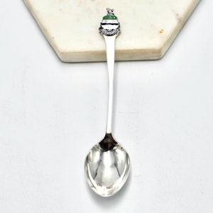 May include: A silver spoon with a decorative handle featuring a green and white design. The spoon is displayed on a white surface, showcasing its polished, reflective bowl and detailed handle.