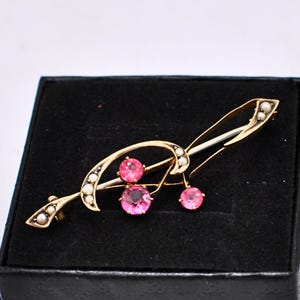 Puede incluir: Broche dorado con un diseño curvo, con tres gemas rosas y pequeñas perlas blancas. El broche se encuentra sobre un fondo negro, mostrando sus elegantes detalles y artesanía.