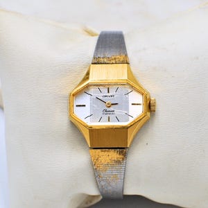 May include: A vintage gold-coloured Orient watch with a rectangular face and a silver-coloured band. The watch face is white with black markers and hands. The watch is resting on a white pillow.