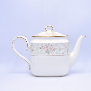 Pode incluir: Bule de porcelana branco com um design floral em rosa, verde e azul. O bule tem uma alça e um bico curvos, e uma tampa com um anel dourado. Uma peça clássica para servir chá.