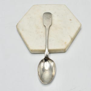 May include: A silver spoon with a rectangular handle and a rounded bowl, resting on a white, hexagonal marble tile. The handle has an engraved design. The spoon is likely vintage or antique.