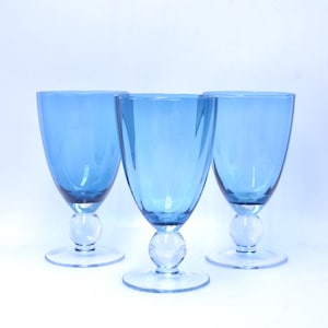 May include: Three matching blue glass goblets with smooth, rounded bowls and clear glass stems featuring a spherical ball accent above a circular base. The glassware has a consistent translucent blue hue and a simple, clean design profile.