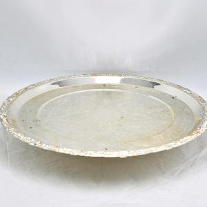 May include: A silver-colored, round serving tray with a decorative, ornate edge. The tray has a slightly raised rim and a smooth, reflective surface. The tray's design includes a subtle, embossed pattern in the center.