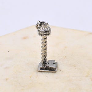 Puede incluir: Un charm plateado con forma de torre estilizada. El charm presenta una sección superior detallada, una columna central en espiral y una base rectangular. El charm está diseñado para ser adjuntado a una pulsera o collar.