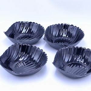 May include: A set of four glossy black bowls, each shaped like a scallop shell with a fluted, ridged exterior and a smooth, curved interior. The bowls are arranged in a two-by-two grid, showcasing their uniform dark finish and shell-like form.