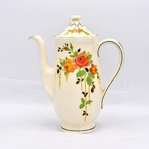 Puede incluir: Cafetera de cerámica color crema con diseño floral. La cafetera presenta un pico largo, un asa curva y una tapa. El diseño floral incluye rosas rojas, naranjas y amarillas con hojas verdes y tallos negros. Ribete negro en la base y el asa.