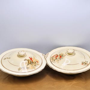 Puede incluir: Dos fuentes de servir color crema con tapas. Cada fuente presenta una ilustración decorativa de personas con abrigos rojos a caballo. Las tapas tienen un pequeño pomo redondo y un borde dorado. Las fuentes están sobre una superficie marrón.