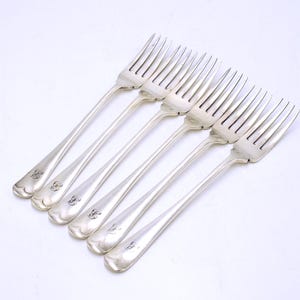 May include: Six silver forks with long, slender handles and tines are arranged diagonally on a white surface. Each fork has a decorative emblem near the base of the handle. The forks are likely for formal dining.