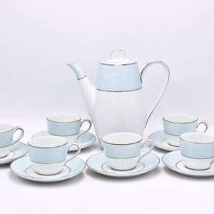 Może przedstawiać: Porcelanowy zestaw do herbaty w kolorze białym i jasnoniebieskim. Zestaw zawiera imbryk, sześć filiżanek i spodków. Imbryk i filiżanki mają jasnoniebieski pasek z kwiatowym wzorem. Srebrne wykończenie.