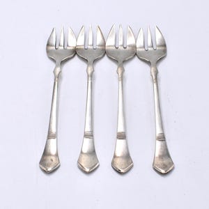 Puede incluir: Cuatro tenedores plateados con tres púas cada uno, dispuestos en fila. Los tenedores tienen mangos largos y delgados con una banda decorativa cerca de la base y un extremo en forma de diamante. Los tenedores son de metal brillante.