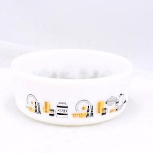 May include: White, round bowl featuring a black and gold kitchen-themed design around the rim. The design includes illustrations of kitchen tools and food items. The bowl is made of a thick, opaque material.