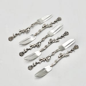 May include: Six silver-toned serving utensils with ornate floral and vine detailing. The set includes three cake servers and three forks, all with a decorative, textured handle. The utensils are arranged diagonally on a white surface.