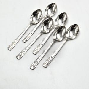 Puede incluir: Seis cucharas plateadas con superficie reflectante dispuestas sobre fondo blanco. Los mangos tienen un diseño floral decorativo. Estas cucharas son adecuadas para servir postres o café.