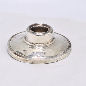 Könnte beinhalten: Ein silberfarbener Kerzenhalter mit breitem, kreisförmigem Sockel und einer zentralen, zylindrischen Tasse. Das Metall hat eine leicht gehämmerten Textur und eine reflektierende Oberfläche, die auf eine Vintage- oder antike Ästhetik hindeutet. Für eine einzelne Kerze.