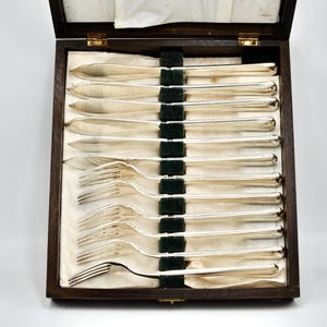 May include: A collection of silver-coloured cutlery, including knives and forks, presented in a wooden box. The box is open, displaying the utensils on a cream-coloured fabric. The silverware is clean and appears to be in good condition.