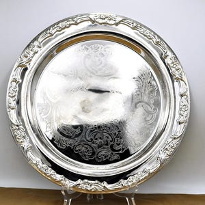 Op de afbeelding: Een ronde, zilverkleurige serveerschaal met sierlijke bloemen- en krulwerkdetails. De schaal heeft een brede, decoratieve rand en een centraal gebied met geëtste patronen. De schaal wordt op een transparante standaard gepresenteerd.